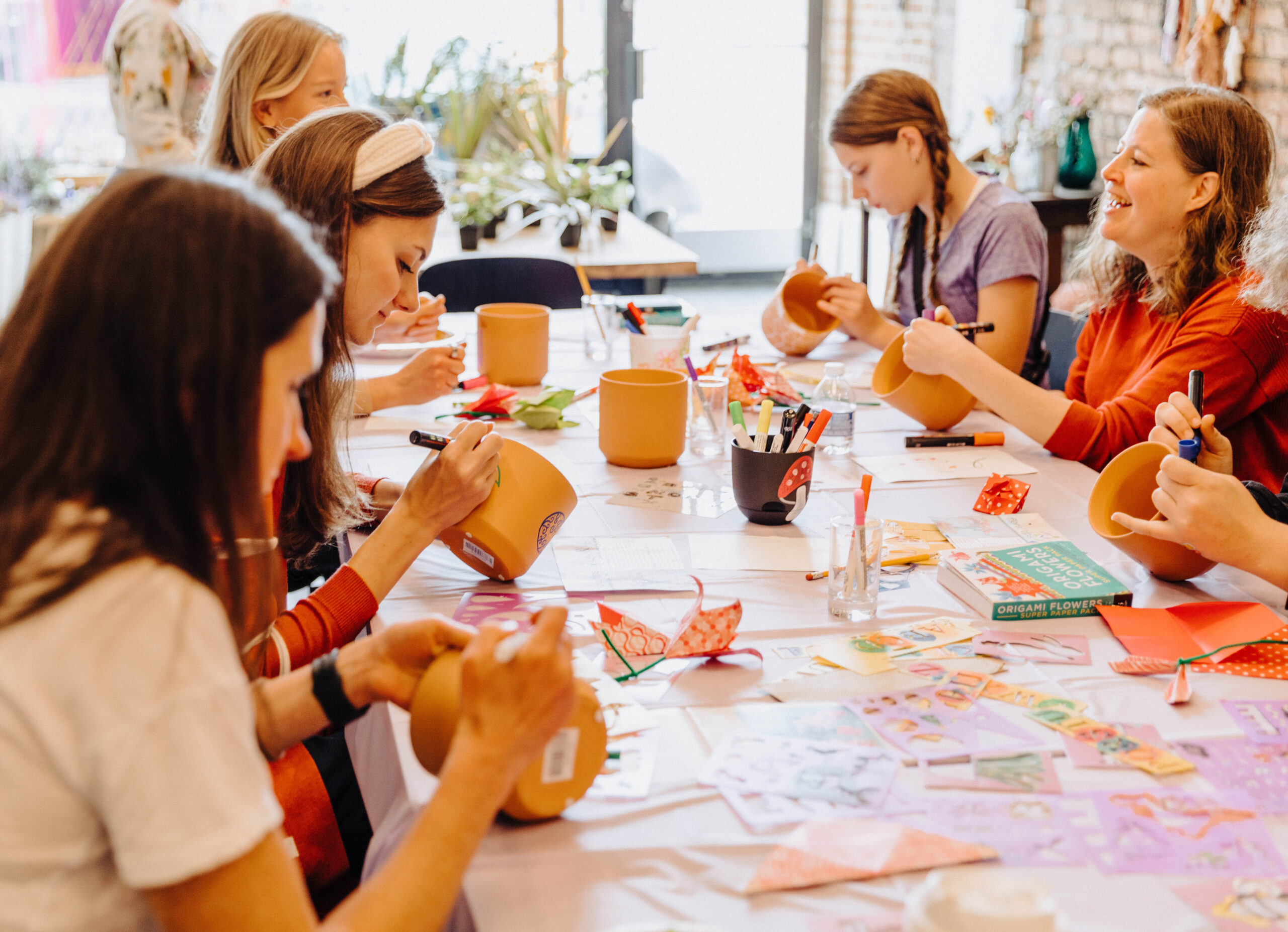 A group of people sitting at a table, painting terracotta pots with markers. The table is covered with craft supplies like paper, markers, and scissors. The setting is bright and cheerful, with large windows and plants in the background.